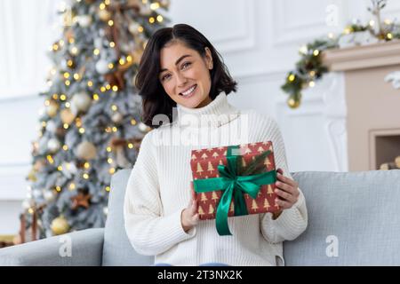 Portrait of a young beautiful woman sitting at home on the couch for New Year's Eve near the Christmas tree, holding a festive gift and smiling at the camera. Stock Photo