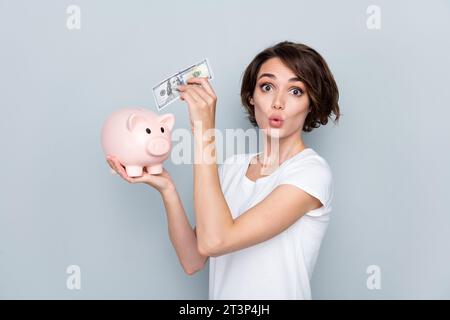 Portrait of funny funky playful girl bob hairdo wear stylish t-shirt put dollar in piggy box plump lips isolated on gray color background Stock Photo