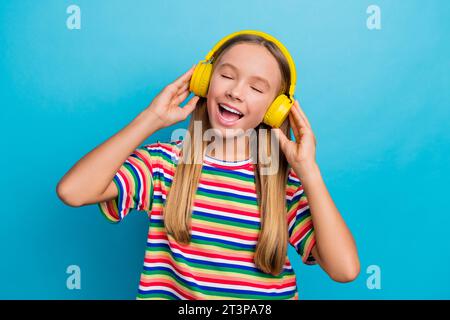 Photo of satisfied funky schoolgirl dressed striped t-shirt touching headphones eyes closed enjoy sound isolated on blue color background Stock Photo