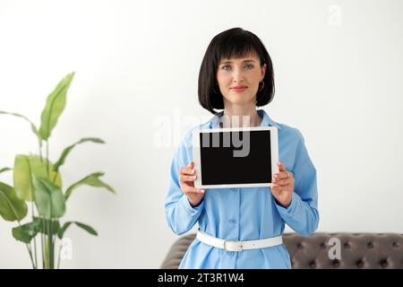 Portrait of young confident modern smiling businesswoman in formal blue shirt shows digital tablet with empty black screen, and looks at camera. Empty Stock Photo