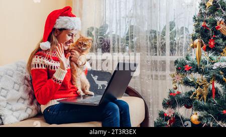 Christmas celebration with cat. Woman in Santa's hat holding pet using laptop by New year tree at home. Calling family in video chat online using inte Stock Photo