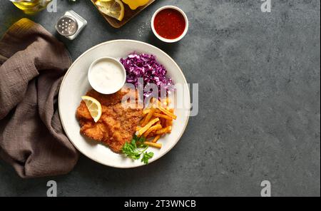 Schnitzel with potato fries, red cabbage salad and sauce on white plate over dark stone background with copy space. Top view, flat lay Stock Photo