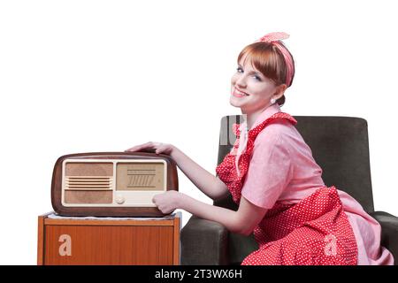 Stereotyped vintage style housewife sitting in the living room and listening to music, she is tuning the radio Stock Photo