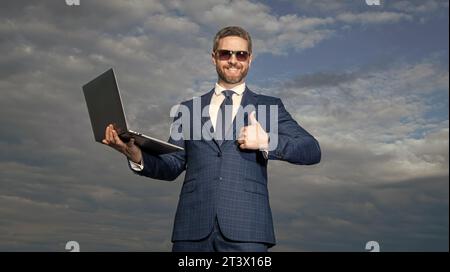 Happy programmer holding laptop. Computer programmer showing thumb gesture. Hardware programmer sky background. Successful business programmer in suit Stock Photo