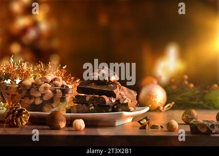 Chocolate with hazelnuts and dried fruit in a white plate on a wooden table decorated in Christmas atmosphere illuminated at night. Front view. Stock Photo