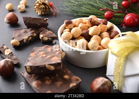 Chocolate with hazelnuts and dried fruit in a bowl on a black table decorated with gift, pine leaves and pine cone at Christmas. Elevated view. Stock Photo
