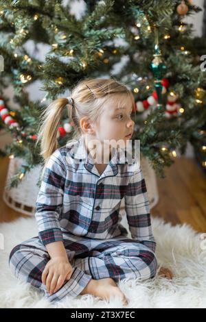 Curious toddler in plaid pajamas sits by Christmas tree Stock Photo