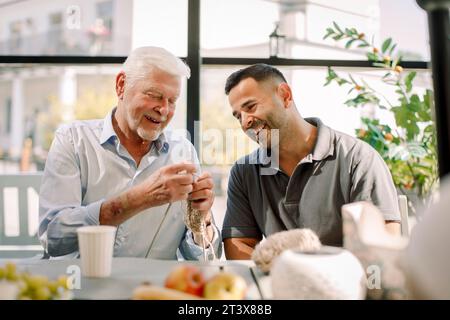 Happy male nurse sitting with senior man crocheting at nursing home Stock Photo