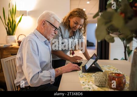 Young female caregiver assisting senior man in using digital tablet at retirement home Stock Photo