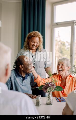Smiling young female caregiver standing by happy senior women and men at dining table in nursing home Stock Photo