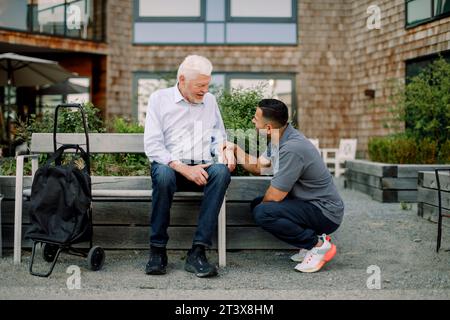 Full length of male nurse squatting while talking to retired senior man sitting on bench at back yard Stock Photo