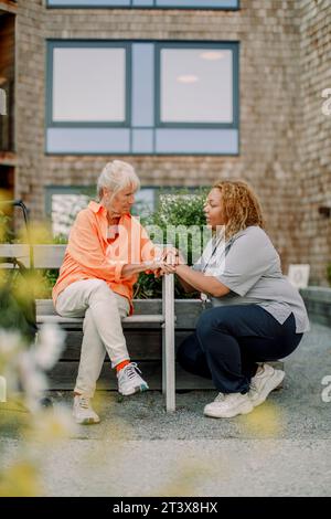 Full length of female nurse squatting while talking to retired senior woman sitting on bench at back yard Stock Photo