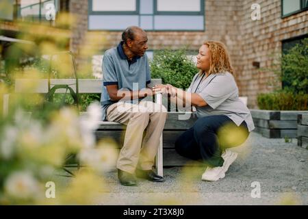 Full length of female nurse squatting while talking to retired senior man sitting on bench at back yard Stock Photo