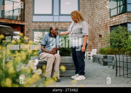 Full length of female nurse with hand on shoulder talking to retired senior man sitting on bench at back yard Stock Photo