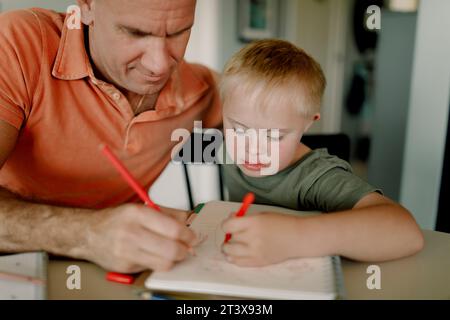 Father and son with down syndrome drawing together in book at dining table Stock Photo