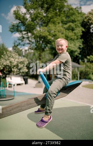 Full length side view portrait of happy boy with down syndrome sitting on seesaw at park Stock Photo