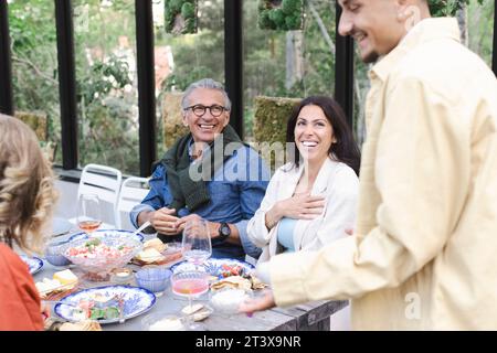 Cheerful family listening to young man talking during dinner party Stock Photo