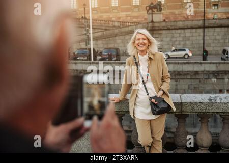 Senior man photographing happy woman leaning on railing Stock Photo