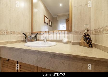 Bathroom tiled with cream-colored marble-like tiles, white porcelain sink on a cabinet with Venetian doors with a mirror built into the wall Stock Photo