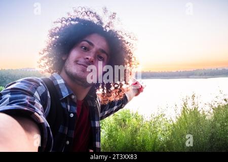 A joyful Arabian man with long curly hair is on a hiking sojourn along a tranquil forest lake trail. With the soft glow of the sunset casting a golden hue, he captures a selfie, his smile radiating the essence of holiday freedom. His backpack, a symbol of adventure, and the serene backdrop of nature come together to create a picturesque frame. Through the lens of his smartphone, he shares a slice of wanderlust, the modern-day explorer reveling in the beauty of the now. Sunset Selfie: Cheerful Arabian Hiker Captures Lakeside Joy. High quality photo Stock Photo