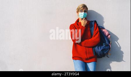 thoughtful woman in glasses and a teal face mask leans against a concrete wall. She wears a red sweater,jeans, and carries a backpack, with one hand r Stock Photo