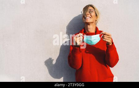 A joyful woman in a bright red sweater removing a light blue face mask, standing against a neutral background, with her shadow visible. Mask freedom a Stock Photo