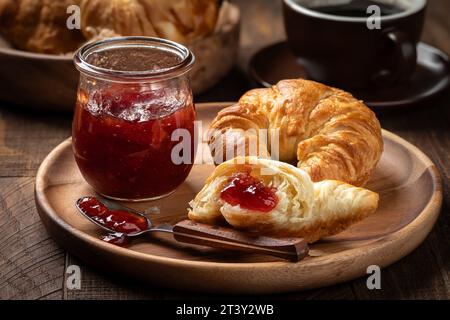 Croissant with strawberry preserves on wooden plate with croissants and cup of coffee in background Stock Photo
