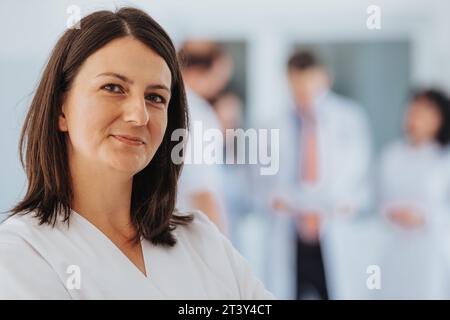Experienced Female Doctor Posing with Confidence, Looking at Camera. Group of Doctors Discussing in The Back. Stock Photo