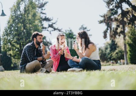 Three carefree friends enjoy a sunny day in a city park, laughing and eating ice cream on the grass. Stock Photo