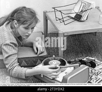 Woman at a gramophone in the 1950s. A typical 1950s teenage girl lying on the floor listening to music from a small gramophone. She is playing the type of records called singles. A record that typically contained a recording of fewer tracks than an LP record. This single record was played at the speed 45 rpm (revolutions per minute) and was 7 inches in diameter, 17,8 cm. The first single record was released 31 march 1949 by RCA Victor. Sweden 1959 Stock Photo
