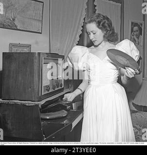 Woman at a gramophone in the 1940s. She's putting another record on the plate to hear.  The records were made of fragile material and the speed of the record to sound as it should was 78 revolutions per minute. A radio is standing on top of the gramophone.   Sweden 1947. Kristoffersson ref V149-3 Stock Photo