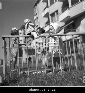 One day in May 1940. A little girl closest to the camera stands is by herself in a playpen with a doll as a toy. A collapsible wooden playpen that has been setup in front of a residential building in central Stockholm. In the background, two slightly older girls are seen playing. Sweden May 1940. Kristoffersson ref 133-3 Stock Photo