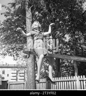 1950s woman. A young blonde woman is seen on a teetering board, in the air, as if the person on the other side is much heavier. She looks happy and smiling. Sweden 1953. Photo Kristoffersson Ref BL96-2 Stock Photo