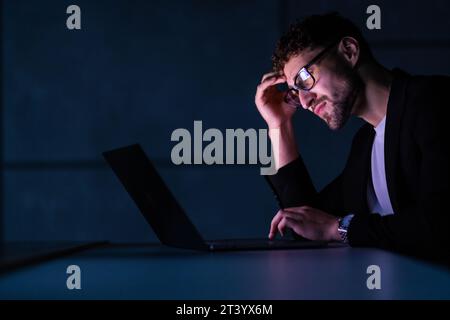 tired male businessman works at home at night on a laptop, works from a home office, studies online,. Internet addiction or a person who works late. a Stock Photo