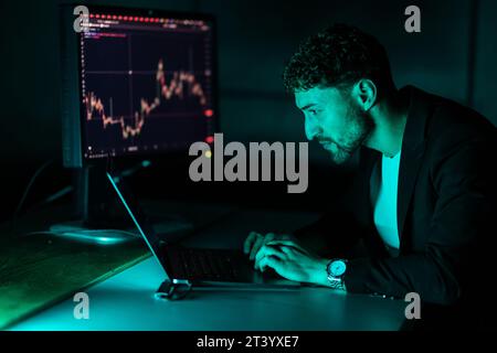 Night portrait of pensive trader looking at laptop monitor, working online with crypto chart in modern office. Online trading, successful business con Stock Photo