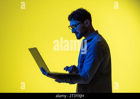 Picture of a pleased young business man posing isolated over light green background wall with led neon lights using laptop computer. Stock Photo