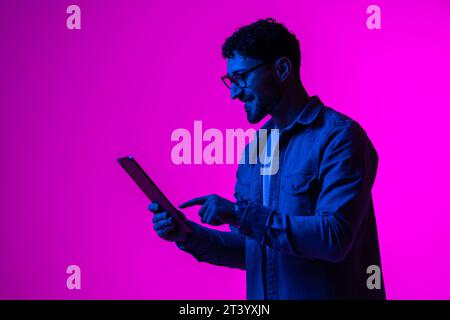Cheerful happy young man using modern digital tablet and wireless stereo headphones, playing video game, watching video content online on red studio b Stock Photo