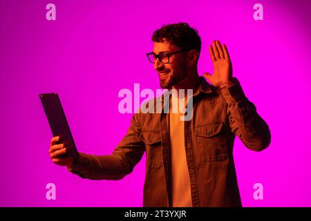 Cheerful happy young man using modern digital tablet and wireless stereo headphones, playing video game, watching video content online on red studio b Stock Photo
