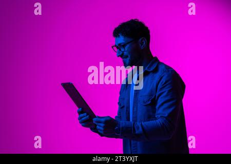 Cheerful happy young man using modern digital tablet and wireless stereo headphones, playing video game, watching video content online on red studio b Stock Photo