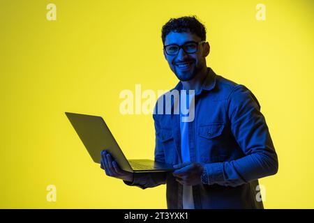 Picture of a pleased young business man posing isolated over light green background wall with led neon lights using laptop computer. Stock Photo