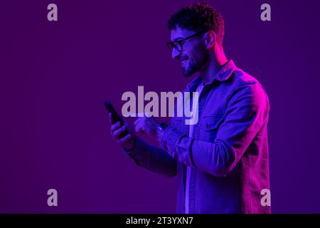 Man smile with phone in hand teenager talking via smartphone online, video call and selfies, hipster lifestyle, portrait purple background, mixed neon Stock Photo