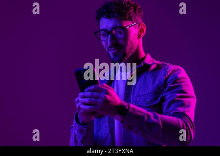 Man smile with phone in hand teenager talking via smartphone online, video call and selfies, hipster lifestyle, portrait purple background, mixed neon Stock Photo