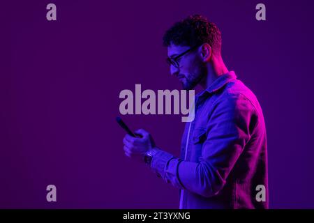 Man smile with phone in hand teenager talking via smartphone online, video call and selfies, hipster lifestyle, portrait purple background, mixed neon Stock Photo