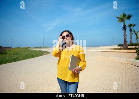 Beautiful young adult woman in sunglasses, standing with a laptop in her hand and looking confidently at camera Stock Photo