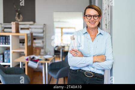 professional smiling tailor woman with hair Braid, wearing glasses and a blue striped shirt, stands in a modern workspace and holding a ruler. Stock Photo