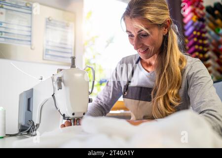 woman in an apron is working at a sewing machine, stitching white fabric in a well-lit workspace with colorful thread spools in the background Stock Photo