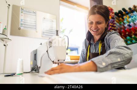 cheerful woman in an apron is working at a sewing machine, stitching white fabric in a well-lit workspace with colorful thread spools in the backgroun Stock Photo