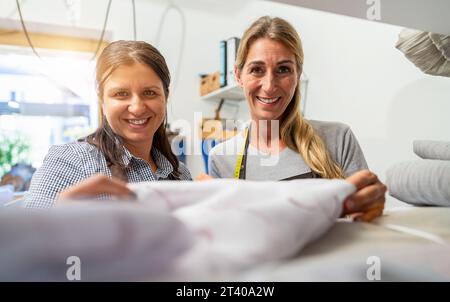 Female work colleagues smiling and closely examining a piece of fabric in a tailoring workshop. One has a measuring tape draped around her neck. Stock Photo