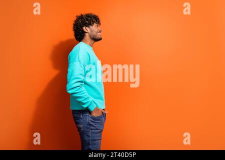 Side shot of young arabian guy wearing stylish bright sweatshirt put hands in jeans look novelty isolated on orange color background Stock Photo
