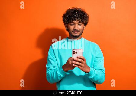 Photo of young confident arabian man wearing teal stylish sweatshirt using smartphone and enjoying app isolated on orange color background Stock Photo